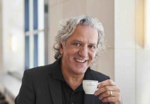 Giorgio Locatelli (soon to be on MasterChef) is seated in a bright interior. He wears a tailored black suit over a black shirt and is holding a small white espresso cup in one hand. His hair is silver and wavy.