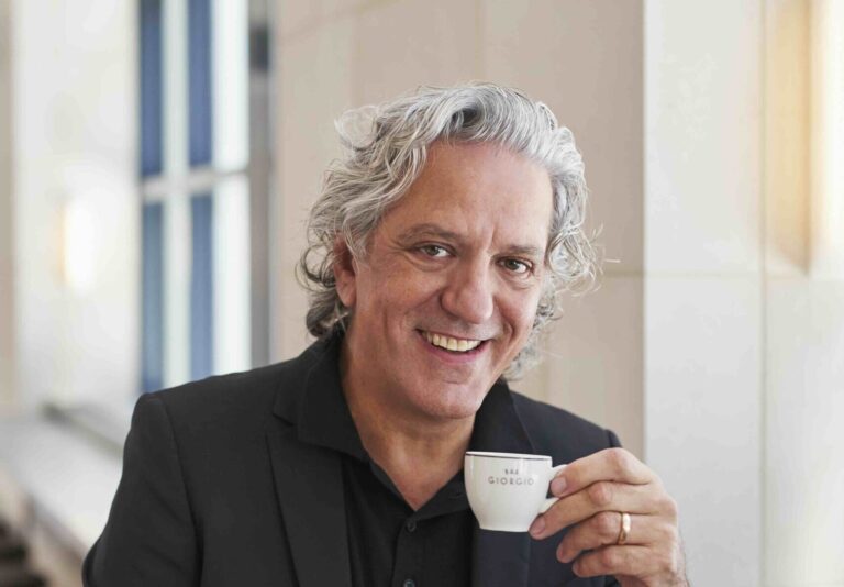 Giorgio Locatelli (soon to be on MasterChef) is seated in a bright interior. He wears a tailored black suit over a black shirt and is holding a small white espresso cup in one hand. His hair is silver and wavy.