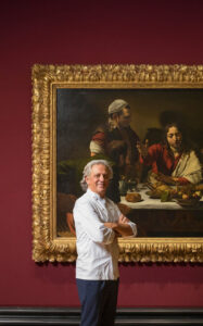 Chef Giorgio Locatelli stands with arms folded in front of a large Caravaggio painting. The artwork, has a large ornate gold frame, depicting a dramatic dinner scene. Hung on a deep red wall.