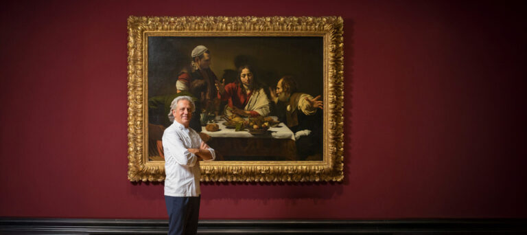 Chef Giorgio Locatelli stands with arms folded in front of a large Caravaggio painting. The artwork, has a large ornate gold frame, depicting a dramatic dinner scene. Hung on a deep red wall.