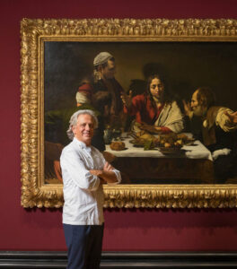 Chef Giorgio Locatelli stands with arms folded in front of a large Caravaggio painting. The artwork, has a large ornate gold frame, depicting a dramatic dinner scene. Hung on a deep red wall.