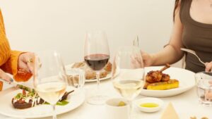 A close-up photograph of a table with three people seated, partially visible, enjoying dishes including steak, roast chicken, and gourmet sides. The table features several glasses of white and red wine, water glasses, and small bowls of olive oil. White background and table cloth.