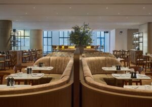 Interior shot featuring beige velvet, curved banquette seating. The space is illuminated by spot lighting on the ceiling. Large windows in the background. The tables are neatly set with white napkins, glassware, and cutlery. There is also some greenery in a vase in the centre of the restaurant.