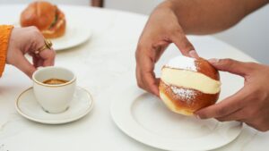 Close up view of a table featuring two people. One hand is about to lift a single cup of cappuccino. The other person is holding a maritozzo, generously filled with cream. There is also a similar bun blurred in the background. The focus is on the hands, the cappuccino, and the creamy filling of the pastry.