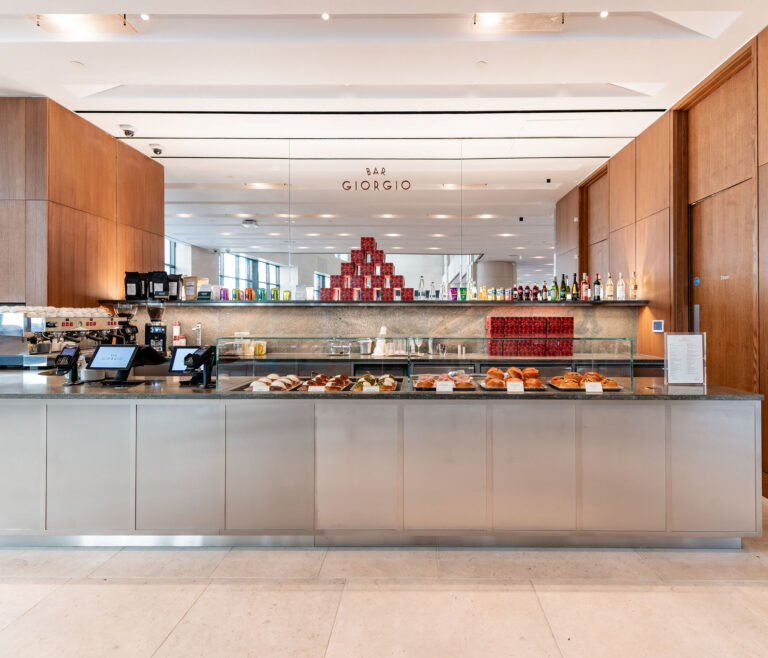 A café counter at Bar Giorgio, featuring an array of pastries displayed behind glass, coffee machines and payment terminals on the left, and shelves of colourful canned goods and bottles of spirits and wine on the mirrored back wall, all set in a bright interior with wood paneling.